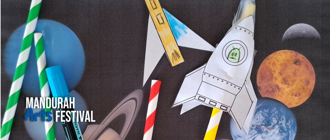 Handmade paper rockets, textas and paper straws are arranged against a backdrop showing planets in outer space